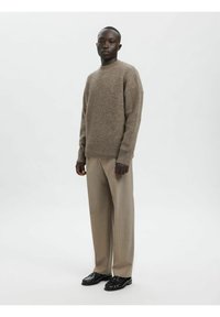 Brūns adīts džemperis ar apaļu kakla izgriezumu, kopā ar gaiši bēšām biksēm un melniem oficiāliem apaviem, stāvot pret vienkāršu fonu.