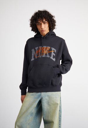 Sudadera negra de Nike con letras grises y logo naranja. Cuenta con un bolsillo frontal y capucha ajustable con cordón. Combinada con jeans azul claro.