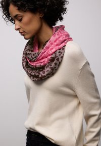 Femme aux cheveux bouclés portant un pull crème et une écharpe infinie à imprimé léopard rose et marron, regardant vers le bas sur un fond uni.