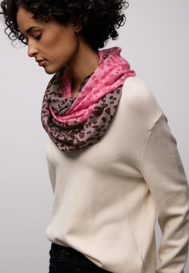 Femme aux cheveux bouclés portant un pull crème et une écharpe infinie à imprimé léopard rose et marron, regardant vers le bas sur un fond uni.