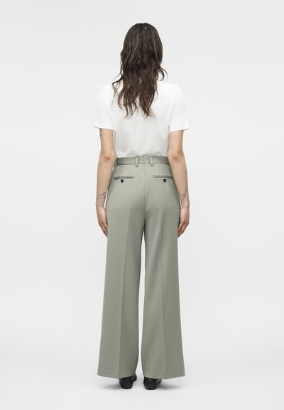 Γυναίκα όρθια με πλάτη στην κάμερα, φοράει λευκό τοπ με κοντά μανίκια και ψηλόμεσα, φαρδιά ανοιχτό γκρι παντελόνια με μαύρα παπούτσια σε απλό φόντο.