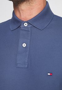 Námořnická modrá polokošile z texturovaného materiálu s tříknoflíkovou légou, bílými knoflíky a malým červeným, bílým a modrým logem na hrudi.