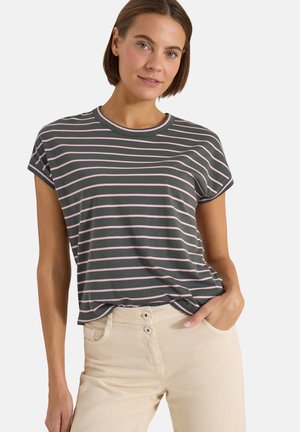 Frau trägt ein dunkelgraues und pink gestreiftes Kurzarmshirt, das in eine hellbeige Hose mit hoher Taille und zwei Knöpfen gesteckt ist, Hand in der Tasche.