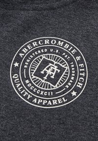 Tessuto grigio scuro con un logo bianco circolare che presenta "Abercrombie & Fitch", un design a forma di libro e testo all'interno di un bordo.