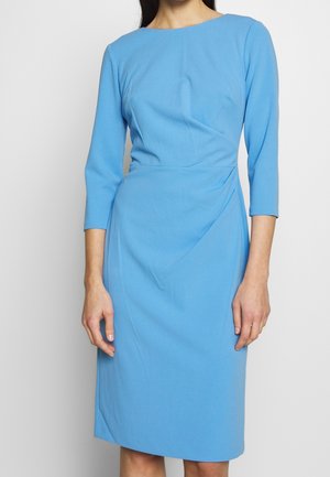 Kobieta ubrana w dopasowaną, sięgającą kolan, błękitną sukienkę z długim rękawem, z subtelnymi plisami i dekoltem typu łódka, stojąca na jednolitym tle.