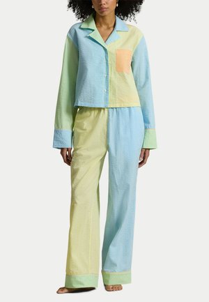 Kobieta ubrana w pastelowy zestaw piżamowy z seersucker, składający się z koszuli na guziki z długim rękawem oraz szerokich spodni w odcieniach niebieskiego, żółtego, zielonego i pomarańczowego.