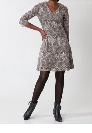 Frau trägt ein knielanges, gemustertes V-Ausschnitt-Kleid mit Dreiviertelärmeln, schwarze Strumpfhosen und schwarze Stiefeletten, posiert mit einer Hand in der Hüfte.