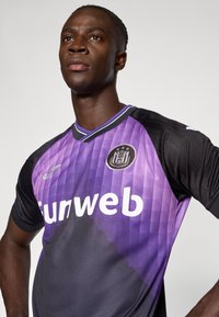 Μωβ και μαύρη φανέλα ποδοσφαίρου με διαμαντένιο μοτίβο, κοντά μανίκια, στρογγυλή λαιμόκοψη και λογότυπο στο στήθος. Λεπτομέρειες σε λευκό.