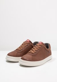 Zapatillas deportivas de cuero marrón con un acabado suave, que presentan cordones beige, una suela intermedia blanca y sutiles rayas verticales en los laterales. Marca roja en la lengüeta.