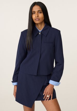 Vrouw met lang, steil haar die een marineblauw colbert met krijtstrepen draagt met een puntige kraag en een bijpassende asymmetrische rok, staand tegen een effen achtergrond.