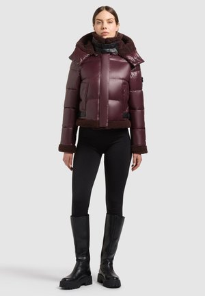 Femme portant une veste matelassée violet foncé avec bordure en fourrure, des leggings noirs et de hautes bottes noires, debout devant un fond uni.