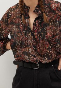 Blouse noire en voile avec un imprimé floral rouge et or, comportant un col à volants et des manches amples, associée à une ceinture noire sur un pantalon sombre.