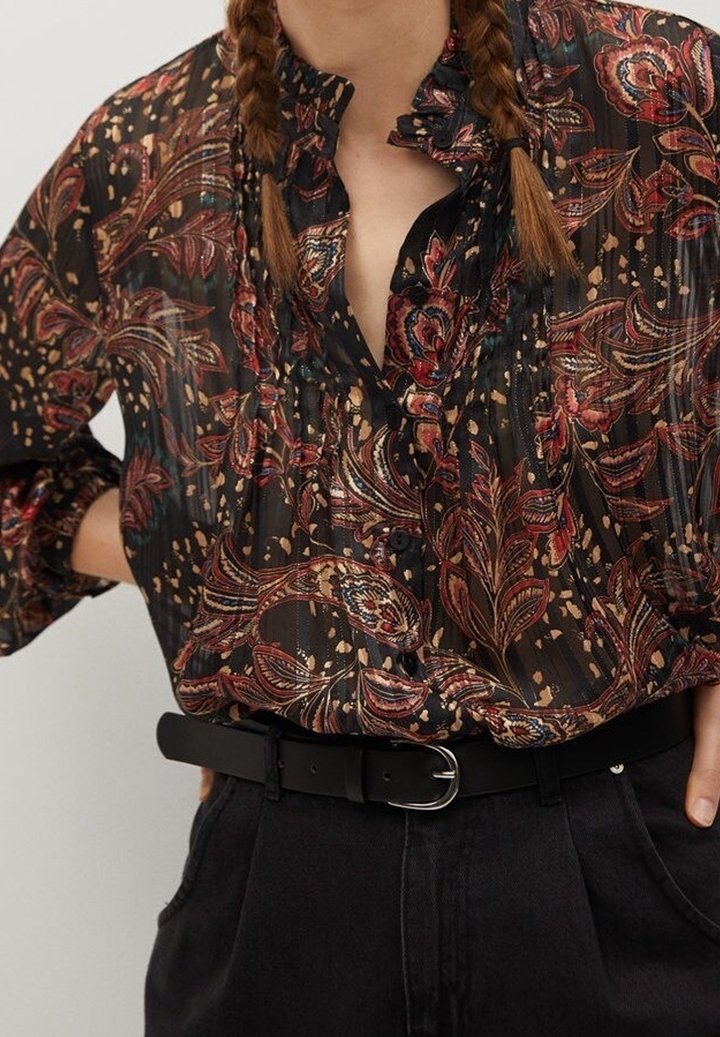 Blouse noire en voile avec un imprimé floral rouge et or, comportant un col à volants et des manches amples, associée à une ceinture noire sur un pantalon sombre.