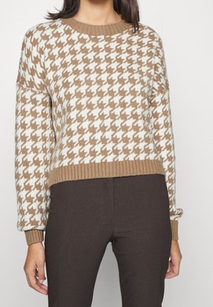 Vrouw draagt een beige en wit trui met een ruitmotief, geribbelde manchetten en zoom, gecombineerd met donkerbruine broek.