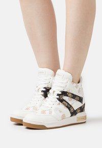 Białe sneakersy typu high-top z złotym wzorem logo, czarnymi akcentami i teksturowaną gumową podeszwą. Związane z przodu.