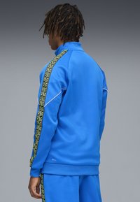 Sininen urheilu takki, joka on valmistettu sileästä kankaasta, ja jossa on vihreä geometrinen kuviointi sivussa ja korkea kaulus.