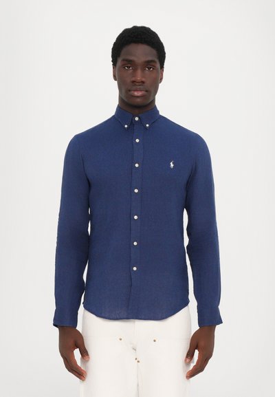 Marineblå knappskjorte laget av teksturert stoff, med en liten logo på brystet, hvite knapper og krage.