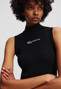 Čierny rebrovaný bezrukávový top s vysokým golierom, na prednej strane s bielym vyšívaným nápisom "KARL LAGERFELD JEANS". Hladká textúra.