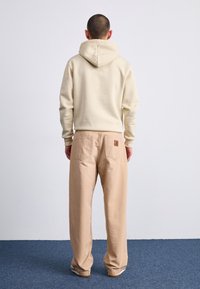 Beige hoodie med snøre i hætten, iført over lyse beige løst siddende bukser. Fremtrædende lommedesigns på bukserne. Glat tekstur.