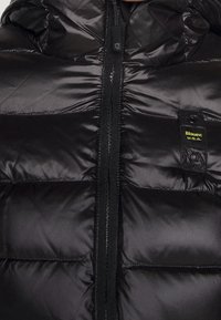 Czarna, błyszcząca kurtka puchowa z centralnym zamkiem błyskawicznym i małą naszywką na piersi z napisem "Blauer: U.S.A." w żółtym kolorze.