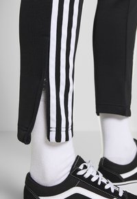 Černé sportovní kalhoty s bílými pruhy po stranách, s zipem na kotnících. V kombinaci s černými teniskami a bílými ponožkami.