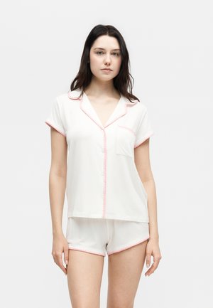 Γυναίκα που στέκεται, φοράει λευκό παντελόνι πιτζάμας με κοντά μανίκια και αντίστοιχα σορτς με ροζ περιθώριο, ουδέτερη έκφραση, απλό φόντο.