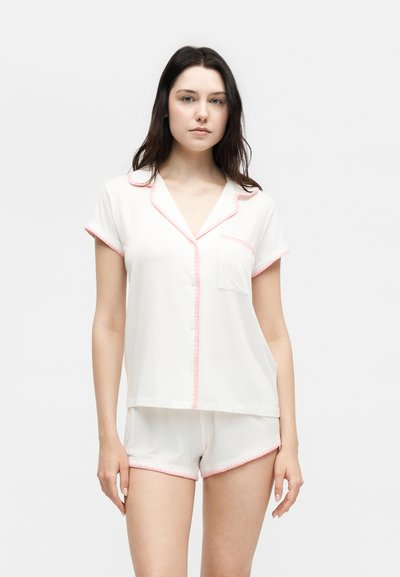 Femme debout portant un haut de pyjama blanc à manches courtes et un short assorti avec un bord rose, expression neutre, fond uni.