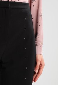 Svarta byxor med en slät textur, försedd med metallnitar längs sidan; bärs med en ljusrosa blus med svarta stjärnmönster.