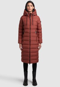 Long, puffy coat in rust orange, featuring a hood, zip closure, and side pockets with black zippers. Paired with black slim pants and boots.