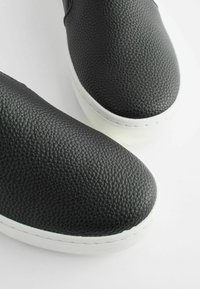 Svart texturerad läderskor med slip-on-design och rund tå, vit gummisula och synlig sömnad längs kanten.