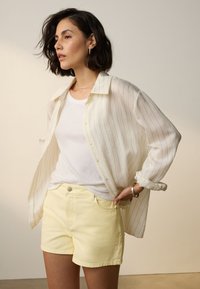 Chemise à rayures blanches boutonnée portée par-dessus un débardeur blanc, associée à un short en denim jaune pastel. Textures lisses, coupe décontractée, accessoires minimaux.