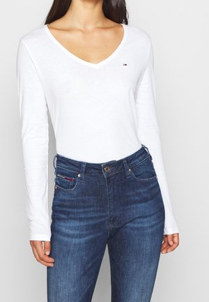 Frau trägt ein weißes Langarmshirt mit V-Ausschnitt, das in hoch taillierte, dunkelblaue, eng anliegende Jeans gesteckt ist.