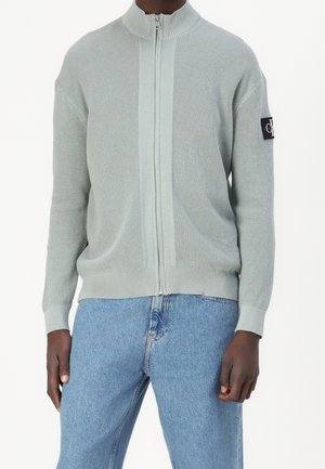 Homme portant un pull zippé en maille gaufrée gris clair et un jean bleu clair, debout avec les bras détendus le long du corps.