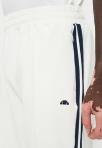 Pantalones deportivos blancos con rayas laterales en negro y blanco y un pequeño logo negro cerca del bolsillo, llevados por una persona con vitiligo en la mano.