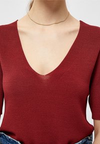 Bordowy sweter z dzianiny o głębokim dekolcie w kształcie V i krótkich rękawach. Posiada teksturowany wzór i luźny krój. Prosta złota naszyjnik choker.