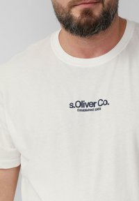 Biała bawełniana koszulka z okrągłym dekoltem i krótkimi rękawami, z granatowym nadrukowanym logo, które brzmi "s.Oliver Co. Założona w 1969 roku."