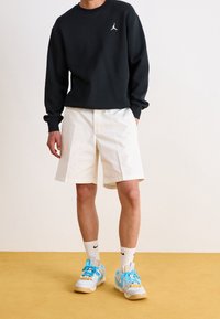 Sudadera negra con un logo blanco, pantalones cortos crema y zapatillas de colores con cordones y detalles azules, calcetines blancos con detalles de logo.