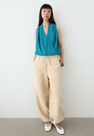 Femme portant un haut cache-cœur sans manches bleu sarcelle, un pantalon large beige, des ballerines blanches texturées et un bracelet en bois, debout contre un mur blanc.