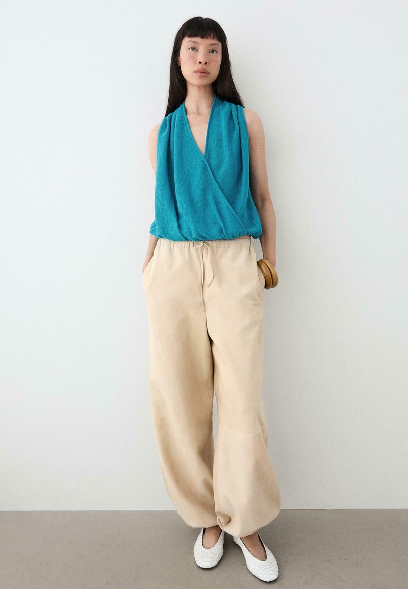 Femme portant un haut cache-cœur sans manches bleu sarcelle, un pantalon large beige, des ballerines blanches texturées et un bracelet en bois, debout contre un mur blanc.