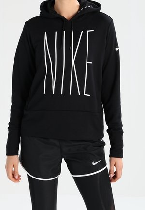 Personne portant un sweat à capuche Nike noir avec un grand logo blanc, un short Nike noir et des leggings noirs sur un fond clair uni.