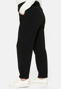 Osoba v černých zúžených kalhotách s elastickými manžetami a bílých teniskách, stojící s jednou rukou v kapse na bílém pozadí.