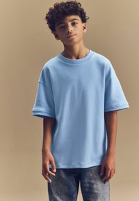 Svijetloplava oversize majica kratkih rukava, izrađena od mekane tkanine; uparena sa svijetlim trapkama, prikazana na neutralnoj pozadini.