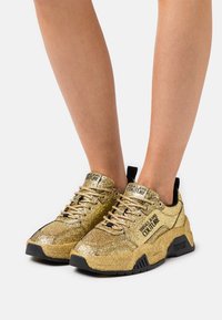 Guldglittriga sneakers med svarta accenter, featuring en tjock sula, rund tå och snördesign. Logotyp synlig på sidorna och tungan.