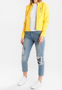 Giacca gialla in ecopelle con tasche con zip, abbinata a jeans azzurri chiari strappati con toppe e sneakers bianche. Outfit casual.