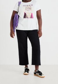 En person som bär en vit T-shirt med en krönt kattgrafik och texten "QUEEN", svarta byxor, svartvita sneakers och bär en lila fransväska.