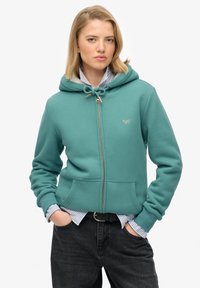 Sudadera con cremallera en color azul verdoso, con capucha ajustable, bolsillos frontales y un discreto detalle de logo. Lleva una camisa a rayas azul y blanca debajo.