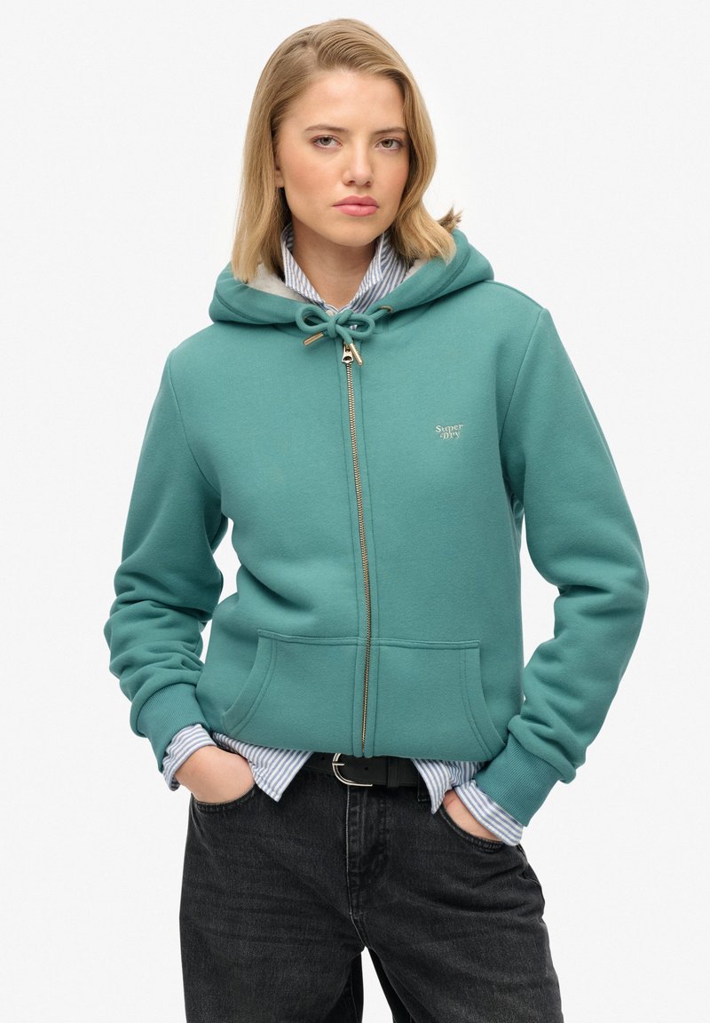 Sudadera con cremallera en color azul verdoso, con capucha ajustable, bolsillos frontales y un discreto detalle de logo. Lleva una camisa a rayas azul y blanca debajo.