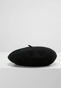 Boina de lã preta com uma forma arredondada, acabamento texturizado e uma pequena alça de tecido no topo, apresentada sobre um fundo neutro.