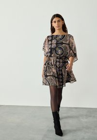 Zwarte jurk met patroon en paisley-design in roze, blauw en geel. Met vloeiende, korte mouwen, elastische tailleband, gecombineerd met zwarte hoge hakken.