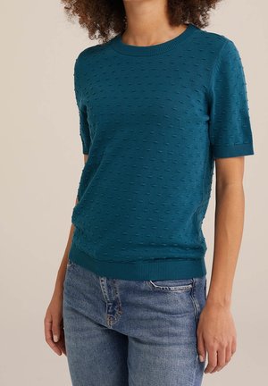 Kvinde iført en teal kortærmet tekstureret strikbluse kombineret med blå denimjeans, stående mod en neutral baggrund.